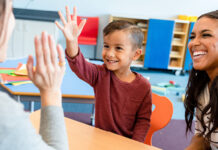 Boy raising hand in classroom with mom and teacher
