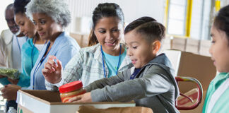 Mid adult mom and her elementary age son sort food items received during a food drive. The little boy is placing a jar of peanut butter into a donation box.